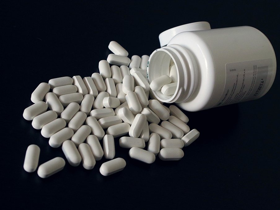 Cápsulas blancas saliendo de un frasco de medicamento sobre fondo oscuro.